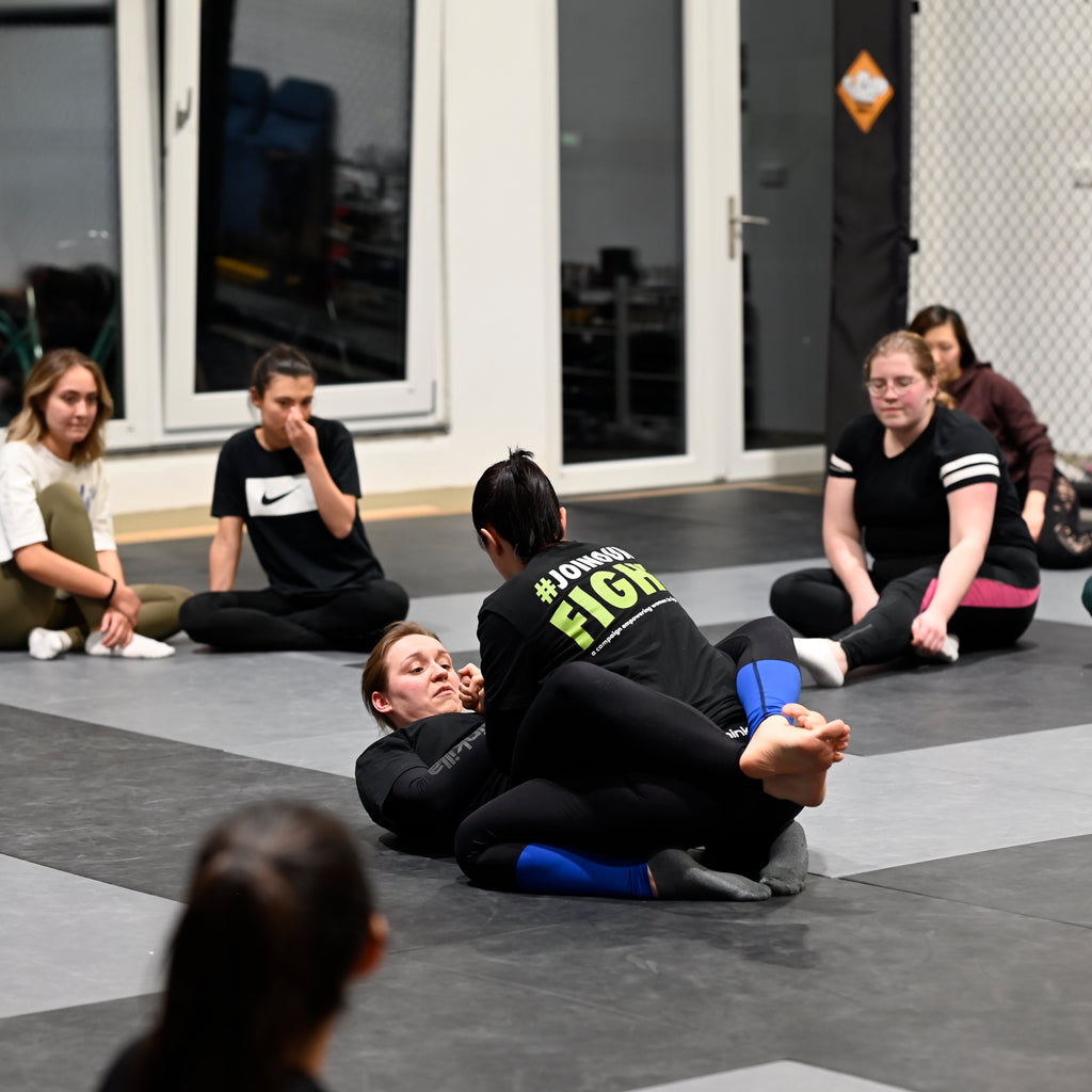 Zwei Kampfsportlerinnen beim Grappling umringt von Frauen. Sie tragen Kampfsportkleidung von Chinkilla. Two female martial artists grapple surrounded by a group of women. They are wearing fight gear by Chinkilla.