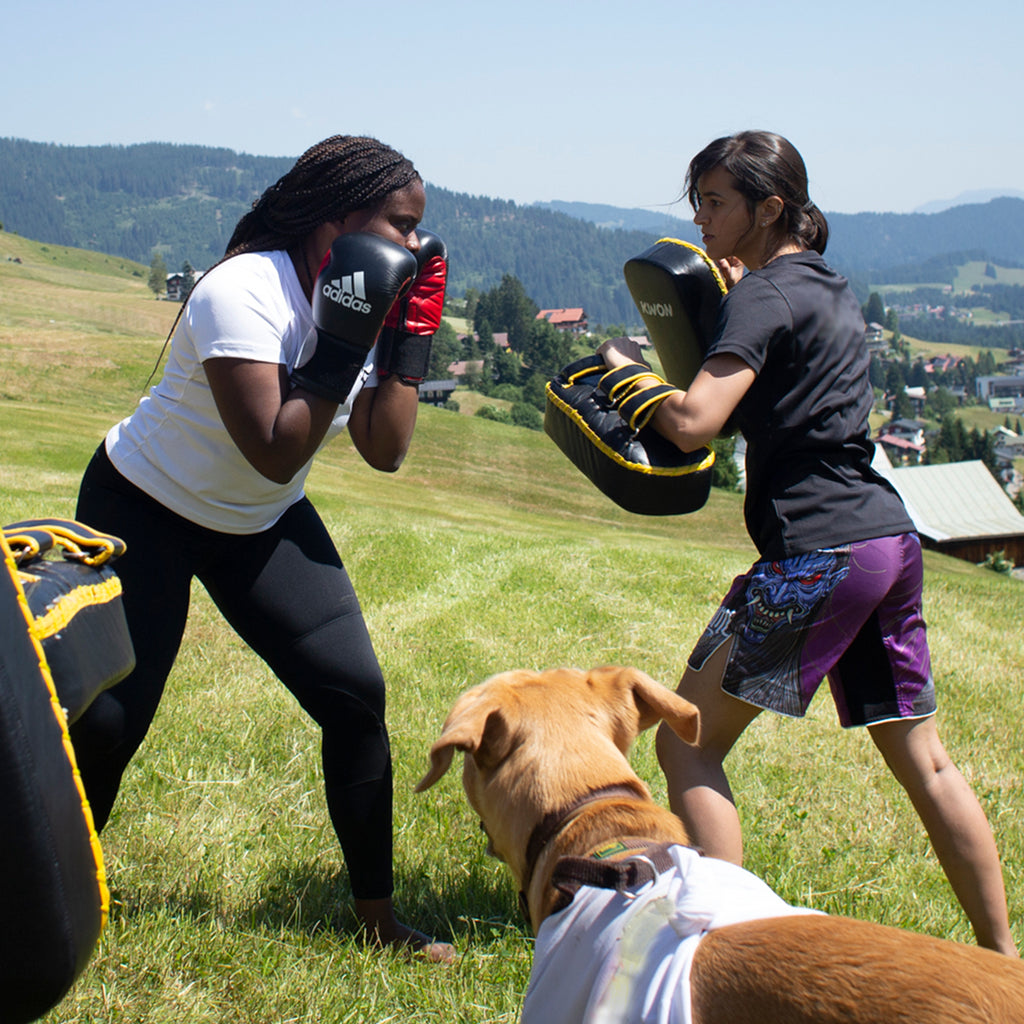 Zwei Frauen beim Kickboxtraining auf einer Wiese in den Bergen während einem Kampfsport Camp der Marke Chinkilla. Two women kickboxing on a field in the mountains during a martial arts camp from the martial arts brand Chinkilla.